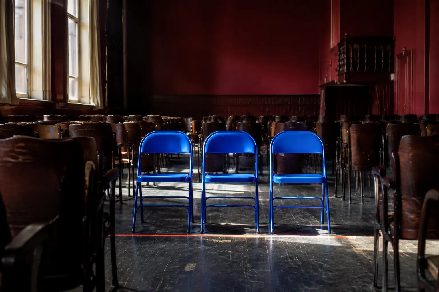 Three vivid cobalt chairs newly placed among rows of older dark chairs in a burgundy rehearsal hall